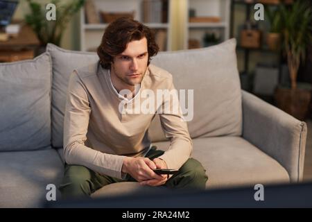 Porträt eines jungen Mannes, der zu Hause fernsieht und auf dem Sofa in einem gemütlichen grünen Innenraum sitzt, Kopierbereich Stockfoto