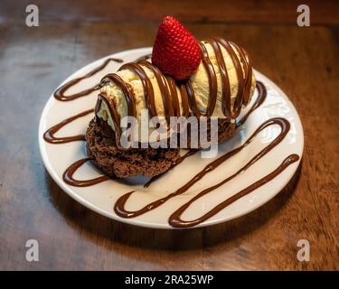 Horizontales Foto eines süßen Brownie-Burgers mit Vanilleeis und Erdbeere, serviert auf einem weißen Teller auf Holzhintergrund Stockfoto