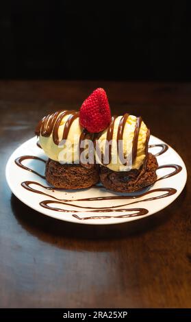 Vertikales Foto eines süßen Brownie-Burgers mit Vanilleeis und Erdbeere, serviert auf einem weißen Teller auf Holzhintergrund Stockfoto