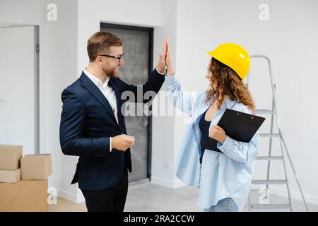 Designerin im Helm mit Dokumenten, die Schlüssel von der Wohnung an einen jungen Geschäftsmann geben, während er im Zimmer steht. Stockfoto