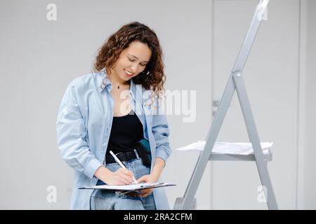 Eine junge, hübsche Designerin bewahrt Dokumente auf, während sie an ihrem Arbeitsplatz steht. Apartment-Renovierung und Designkonzept. Stockfoto