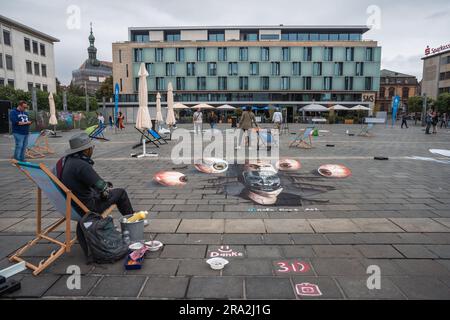 Kaiserslautern, Deutschland. 30. Juni 2023. Der mexikanische Künstler Rafa Rars sitzt auf einem Stuhl vor seinem fertigen Gemälde. Elf internationale Künstler aus 6 Ländern verwandelten den Stiftsplatz (Platz) in dreidimensionale Kunst, wobei das Thema „Digitalisierung und intelligente Stadt“ dieses Jahr stand. 3D Street Art nutzt die Perspektive, um eine optische Illusion des Weltraums zu erzeugen. Die Besucher konnten alle Künstler bei der Arbeit drei Tage lang beobachten. Dies ist der dritte und letzte Tag der Veranstaltung. Kredit: Gustav Zygmund/Alamy News Stockfoto