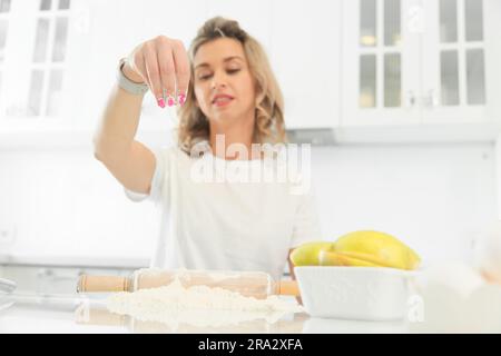 Ein junges Mädchen sitzt an einem Tisch in einer hellen weißen Küche und bereitet ein Mittagessen oder ein Gebäck-Dessert zu. Helle Farbtöne. Eine schöne weiße Blondine schüttet Mehl auf den Tisch. Vorderansicht mit Kopierbereich Stockfoto