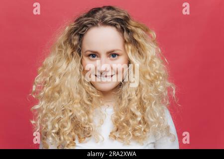 Entzückende junge Frau mit welligen Haaren und blauen Augen. Beißt Lippen, sieht glücklich aus. Rosafarbener Hintergrund Stockfoto