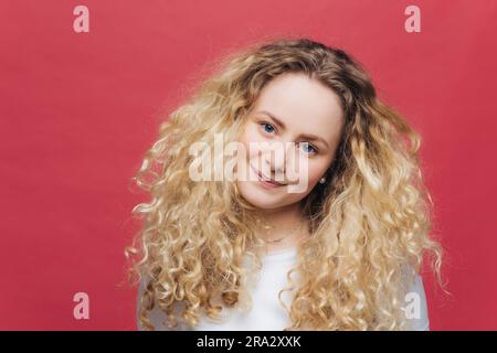 Attraktive Frau mit lockigem Haar und blauen Augen. Positiver Blick, zufrieden. Rosafarbener Hintergrund Stockfoto
