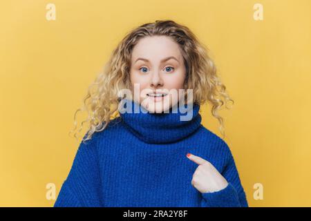 Überraschtes weibliches Model mit lockigem Haar zeigt auf einen leeren Platz auf ihrem neuen Winterpullover, perfekt für Ihre Anzeige oder Ihr Design, auf gelbem Hintergrund. Stockfoto