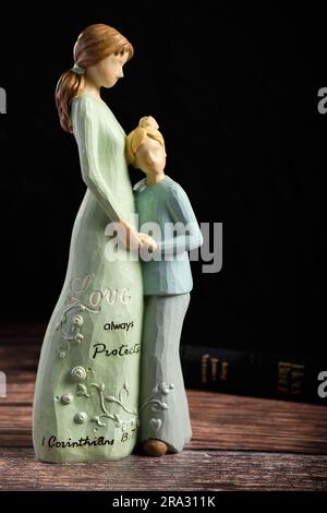 Christliche Keramikdekoration einer Mutter und eines Sohnes mit der Inschrift „Love always protect 1 Corinthians 13:7" vor schwarzem Hintergrund. Stockfoto