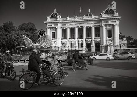 Cyclos (Pedicabs) mit Touristenpässen und anderen Verkehrspässen vor dem französischen Opernhaus aus der Kolonialzeit Hanoi oder Nha hat Lon in Hanoi, Vietnam. Stockfoto