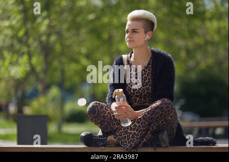 Modische junge Frau in Kleidung mit Leopardenmuster, die auf einer Bank sitzt und eine Glasflasche Wasser in den Händen hält. Porträt eines vielfältigen weiblichen Modells mit Sho Stockfoto