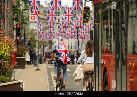 Ein Mann, der mit einer Union Jack Flag verkleidet ist, zeigt seinen Stolz, wenn er auf der britischen Flagge an der Regent Street fährt, die während der Krönungswoche voller Busse ist. Stockfoto