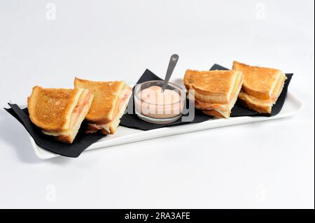 Club Sandwich, in elegante schwarze Servietten eingewickelt, bestehend aus vielen leckeren Zutaten, mit einem Glas Soße, auf weißem Teller und Hintergrund. Stockfoto