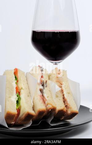 Club-Sandwich auf schwarzem Teller mit einem Glas Rotwein vor weißem Hintergrund. Stockfoto
