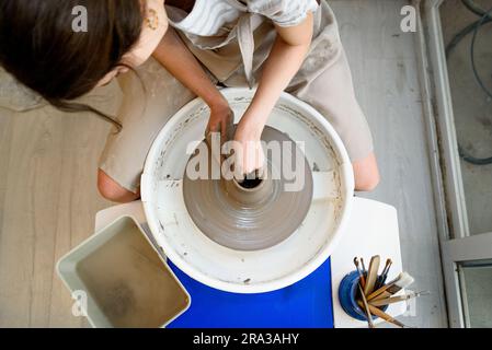 Draufsicht der Drehmaschine mit Händen, die Keramik auf der Drehmaschine oder dem Töpferrad in einer Töpferwerkstatt mit natürlichem Licht bearbeiten und Formen Stockfoto