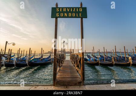 Gondeln, die am Pier mit dem Schild „Gondola Service“ bei Sonnenaufgang auf dem Kanal nahe dem Markusdom in Venedig, Italien, verankert sind. Diese italienische Skyline von Venedig ist unglaublich. Stockfoto