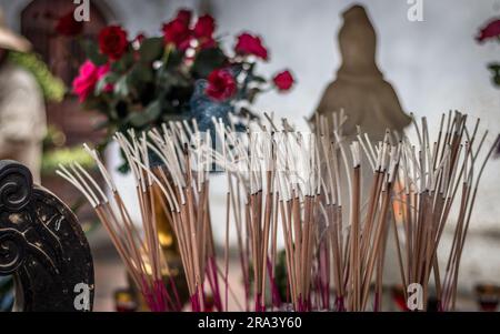 Eine Inneneinrichtung mit verschiedenen aromatischen Räucherstäbchen, lebendigen Blumensträußen und einer Figur an einer neutralen Wand Stockfoto