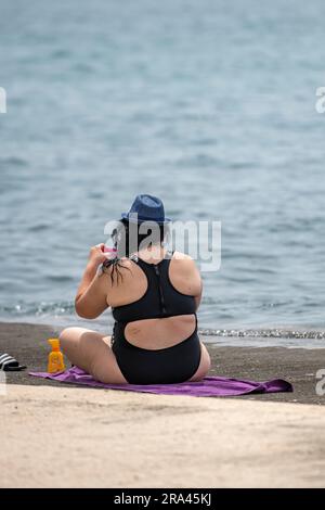 Eine große Dame mit schwarzem Badeanzug und Sonnenhut sitzt am Strand am Wasser. Übergewichtige Frau, die auf einem Handtuch am Meer sitzt. Stockfoto