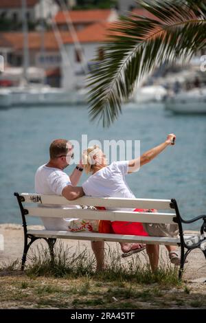 Ein Paar mittleren Alters, das auf einer Bank am Wasser in trogir kroatien sitzt. Ein Paar mittleren Alters, das im Urlaub Selfie-Fotos mit dem Smartphone macht. Stockfoto