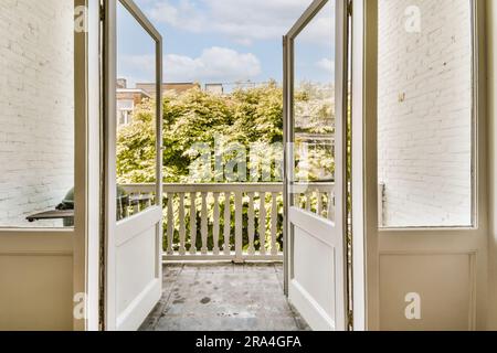 Eine offene Tür führt zu einem Balkon mit weißen Backsteinwänden und grünen Bäumen in der Ferne, an einem sonnigen Tag Stockfoto