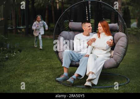 Eine glückliche Familie sitzt in einer Hängematte auf dem Rasen in der Nähe des Hauses. Stockfoto