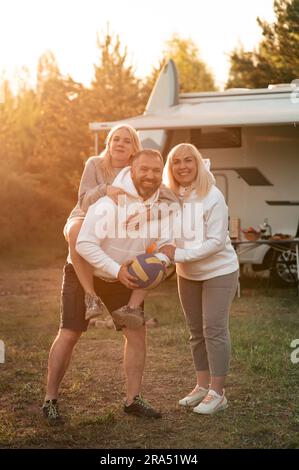 Glückliche Eltern mit ihrem Kind, das mit einem Ball in der Nähe ihres Wohnmobiles im Wald spielt. Stockfoto