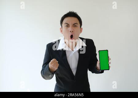 Junger asiatischer Geschäftsmann mit fröhlichem und aufgeregtem Gesichtsausdruck, der ein Smartphone mit grünem Bildschirm hält und es der Kamera zeigt Stockfoto