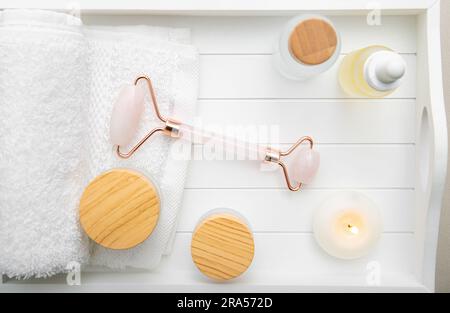 Rosenquarzstein Rollwerkzeug für Gesicht, verschiedene Gesichts- und Körperpflegeprodukte generische Produkte feuchtigkeitsspendende Cremes Öle auf weißem Holztablett. Studiobeleuchtung. Stockfoto