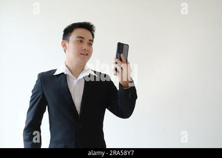 Junger asiatischer Geschäftsmann mit unangenehmem Gesichtsausdruck, der Videoanrufe auf seinem Smartphone macht. Isolierter weißer Hintergrund. Geeignet für Werbung. Stockfoto