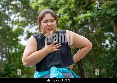 Eine übergewichtige junge Frau mit Brustschmerzen legt ihre rechte Hand über ihre Brustschmerzen während ihres morgendlichen Trainings auf einer Laufstrecke in einem lokalen Park Stockfoto