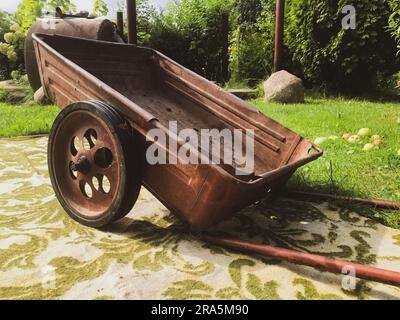 Eine Schubkarre aus Metall mit großen Rädern steht auf einem Teppich. Gartenpflege in russland. Teppich auf der Straße, Mode aus den 90s Jahren. Stilvoll und modisch i Stockfoto