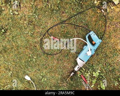 Eine Bohrmaschine mit langem Bohrer liegt auf dem Gras. Renovierung in der Natur. Neben einem Schraubendreher mit rotem Griff. Reparatur eines Holzhauses und -Dachs. Stockfoto