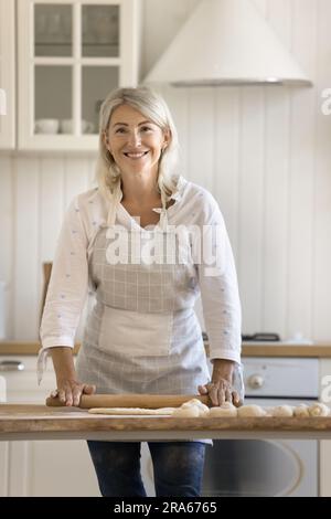 Glückliche, hübsche, blonde, reife Bäckerin mit Schürze und Teig Stockfoto
