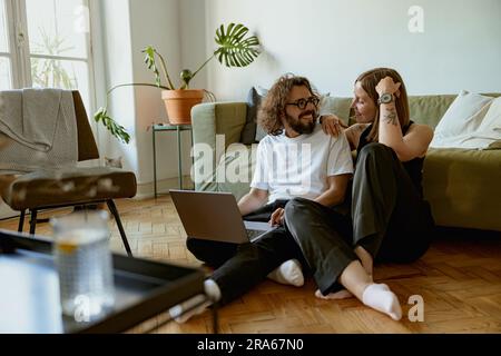 Junge Familie, die sich Videos auf einem Laptop ansieht, sitzt auf dem Boden in einem gemütlichen Apartment. Ein Paar, das sich liebt Stockfoto