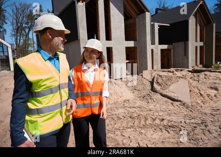 Lächelnde Bauarbeiter, die an den im Bau befindlichen grauen Häusern entlang spazieren Stockfoto