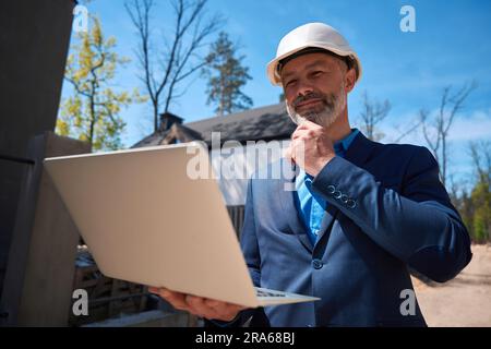 Inspektor im Schutzhelm, der die Baustelle mit App auf dem Laptop überwacht Stockfoto