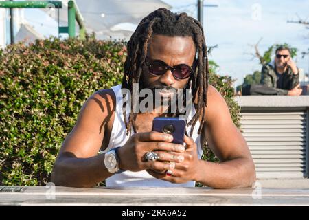 Vorderansicht eines jungen schwarzen Mannes afrikanischer ethnischer Herkunft, mit Dreadlocks und Sonnenbrille, der draußen sitzt, sein Handy aufmerksam beobachtet und die Notiz abfragt Stockfoto