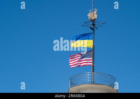 Die Flaggen der Ukraine und der USA flattern auf einem Fahnenmast vor einem blauen Himmel. Statussymbol. Ukrainische blaue und gelbe Flagge. Windtag im Freien. Stockfoto