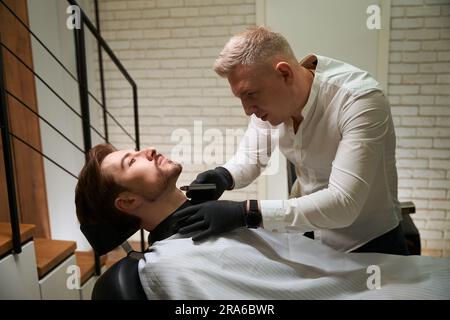 Master rasiert die Bartstoppel am Hals des Kunden mit einem Spezialwerkzeug Stockfoto