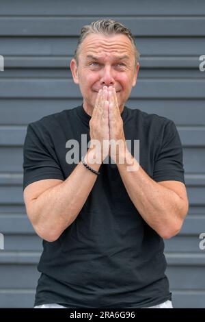 Dreiviertel langes Porträt eines notleidenden älteren Mannes mit beiden Händen am Mund vor einer blauen Wand Stockfoto