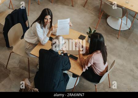 Smiley Brünette zeigt ihr Dokument und stellt eine Frage an ihren 70s erfahrenen Teamleiter. Generationsübergreifende Gruppe von Menschen, die zusammenarbeiten Stockfoto