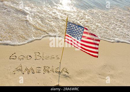 Gott segne Amerika Text im Strand Sand mit einer amerikanischen Flagge auf Sonnenschein Stockfoto