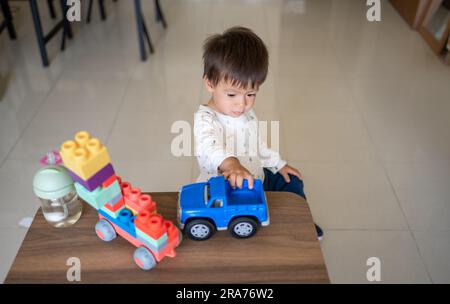 Ein eineinhalb Jahre alter Junge aus gemischten Rassen, der mit bunten Bausteinen und einem formbetonten Spielzeugfahrzeug spielt. Kleinkinder sitzen neben einem Kaffee Stockfoto