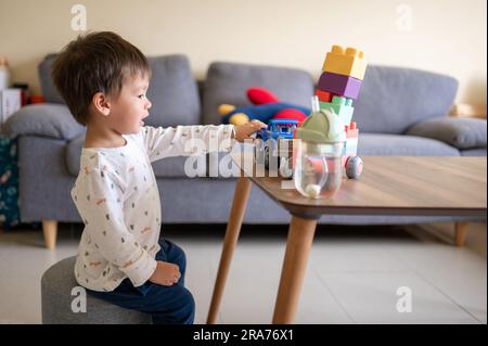 Ein eineinhalb Jahre alter Junge aus gemischten Rassen, der mit bunten Bausteinen und einem formbetonten Spielzeugfahrzeug spielt. Kleinkinder sitzen neben einem Kaffee Stockfoto