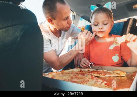 Vater wischte sanft den Mund der Tochter während des Essens gerade gekochte italienische Pizza während einer Familienfahrt mit dem Auto mit Bremsstopp. Glückliche Familienmomente, Kindheit, schnell Stockfoto