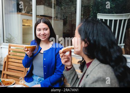 Zwei junge lateinische Geschäftsfrauen, die auf einer bezaubernden Café-Terrasse Kaffee trinken und Erfolg und Freundschaft begrüßen Stockfoto