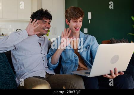 Junge schwule Paare winken und lächeln während eines Videoanrufs auf dem Laptop im Wohnzimmer zu Hause Stockfoto