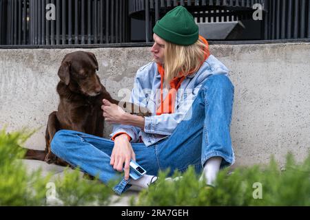 Ein Hipster-Millennials-Typ und ein süßer, obdachloser Hund, der draußen zusammen sitzt Stockfoto