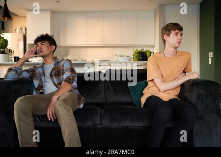 Ein junges schwules Paar in legerer Kleidung, das sich auf dem Sofa im Wohnzimmer zu Hause ignoriert Stockfoto