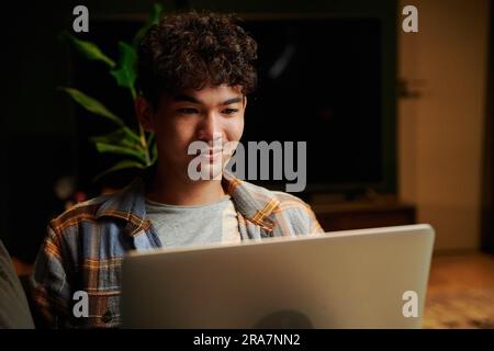Ein fröhlicher junger Multirassenmensch, der ein kariertes Hemd trägt und den Laptop auf dem Sofa im Haus benutzt Stockfoto