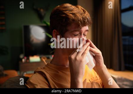 Kranker junger weißer Mann, der zu Hause ein T-Shirt trägt und neben dem Laptop auf dem Sofa Nase bläst Stockfoto