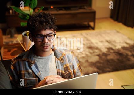 Ein junger, multiethnischer Mann, der eine Brille trägt und zu Hause einen Laptop und kabellose Kopfhörer auf dem Sofa trägt Stockfoto